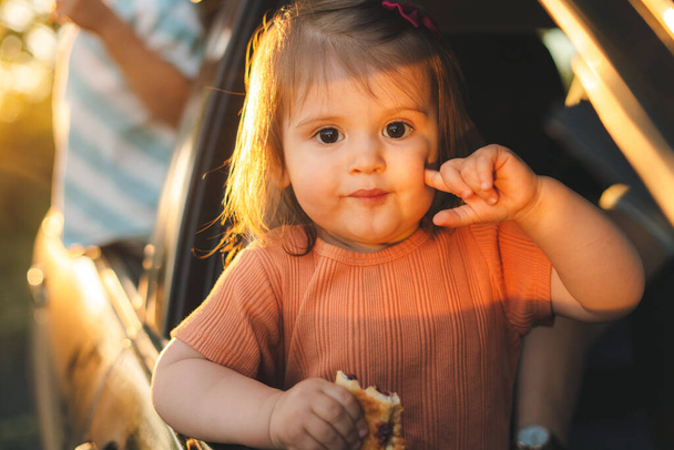 Baby girl in the car looking throw window while eating tasty pie. People looking outside. Childhood concept. - Foto, Imagem