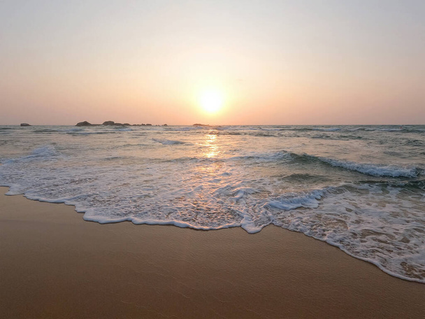 Puha kék óceán hullámok egy tiszta homokos strandon naplementekor. - Fotó, kép