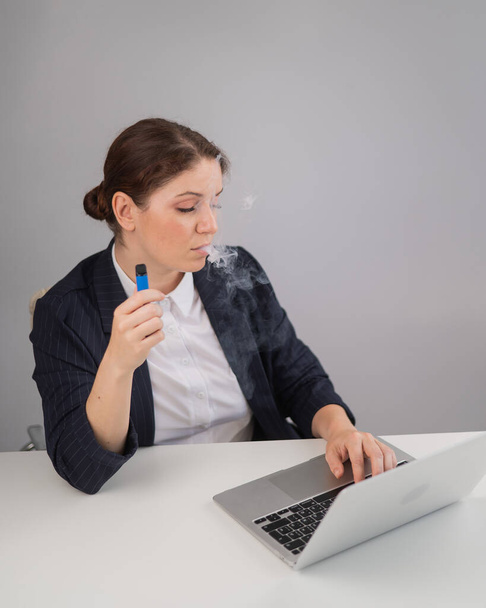 Business woman smoking a disposable vape while sitting at her desk - Photo, image
