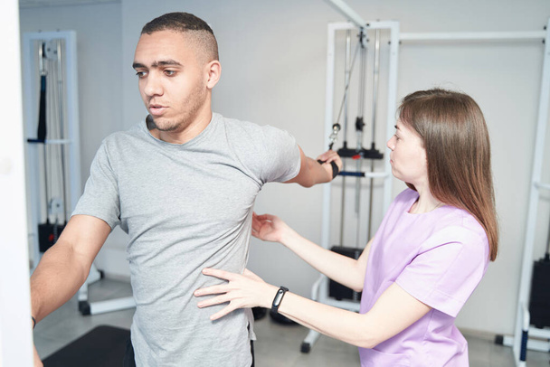 African American man stretching arms and using medical kinesiotherapy equipment while doing physiotherapy exercise with female physiotherapist - Foto, Bild