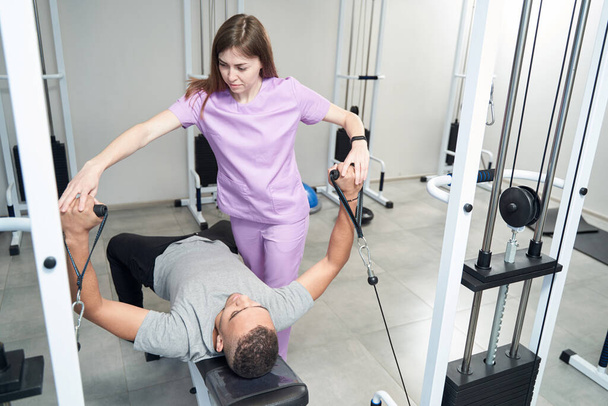 African American man using medical kinesiotherapy equipment while having rehabilitation gymnastics training with female physiotherapist - Foto, Imagem