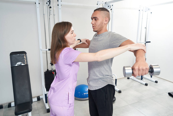 Пациент мужского пола делает физиотерапевтические упражнения с гантелями во время занятий кинезиотерапией с врачом в клинике - Фото, изображение