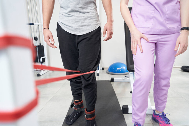Male patient using kinesiotherapy exercise equipment while having rehabilitation training with physiotherapist after leg injury - Photo, Image