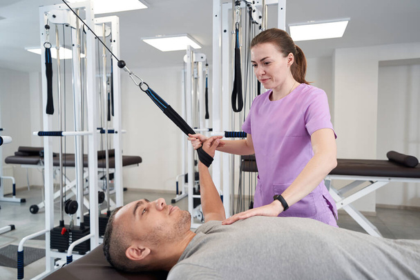 Male patient using kinesiotherapy training equipment while having rehabilitation workout with woman physiotherapist - Photo, image