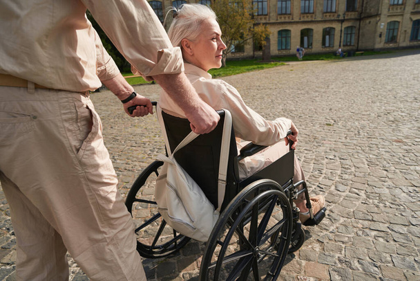 Mature male person standing behind his partner while pushing the wheelchair - Photo, image