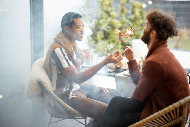 Kaksi miespuolista ystävää polttamassa tähtisadetikkuja takapihalla. Valkoihoinen ja latinomies juhlivat yhdessä. Idea homoparien ja loma tunnelma - Valokuva, kuva