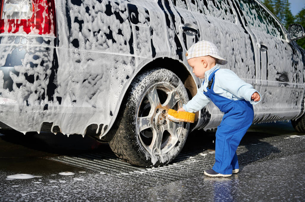 Toddler child leaning over rear wheel of foamy car and holding sponge for washing auto, testing himself in the profession of car washer outdoors in the afternoon. - Photo, Image