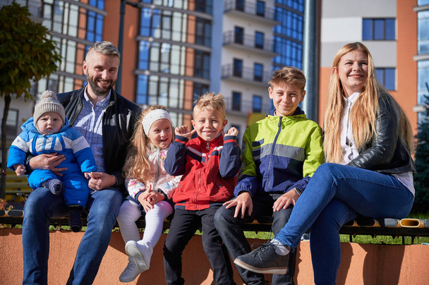 Two adults spending time with cute children, smiling in spring sunny day. Front view of middle-aged parents sitting on bench with four adorable kids, having rest in city centre. Concept of family. - Foto, imagen