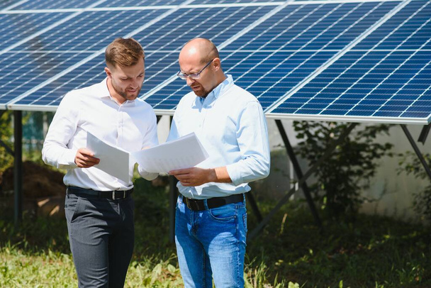 2人のエンジニアと太陽光発電所(太陽光パネル)は、システムの動作を確認するために歩く、世界のエネルギーを節約するための代替エネルギー、クリーンエネルギー生産のための太陽電池モジュールのアイデア. - 写真・画像