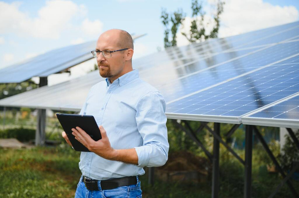 Portrait of male engineer with tablet in his hands near the solar panels station, wearing helmet at sunny day. Green ecological power energy generation - Photo, image