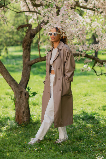 молодая африканская американка в стильных солнцезащитных очках и плаще прогуливаясь с руками в карманах в парке - Фото, изображение