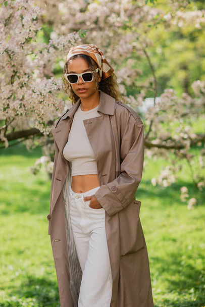 young african american woman in stylish sunglasses and trench coat standing with hand in pocket near blooming tree - 写真・画像
