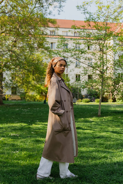 young african american woman in headscarf and stylish trench coat standing with hand in pocket on green grass in park - Valokuva, kuva