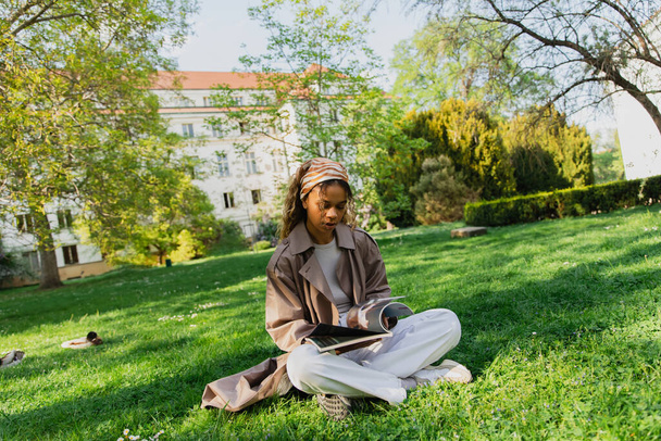 νεαρή Αφροαμερικανή γυναίκα με μαντίλα και κομψή καμπαρντίνα κάθεται στο πράσινο γρασίδι και διαβάζοντας περιοδικό στο πάρκο - Φωτογραφία, εικόνα