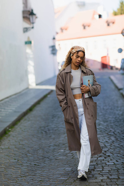 full length of african american woman in headscarf and trench coat walking with magazine on street in prague  - Fotografie, Obrázek