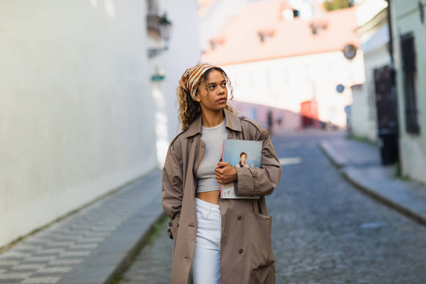 stylish african american woman in headscarf and trench coat walking with magazine on street in prague  - Φωτογραφία, εικόνα