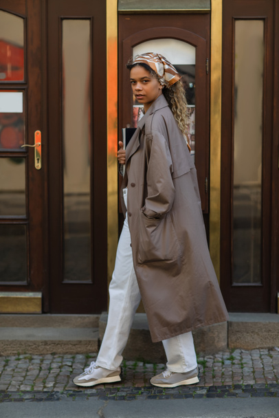 full length of stylish african american woman in headscarf and trench coat walking with hand in pocket in prague - Photo, Image
