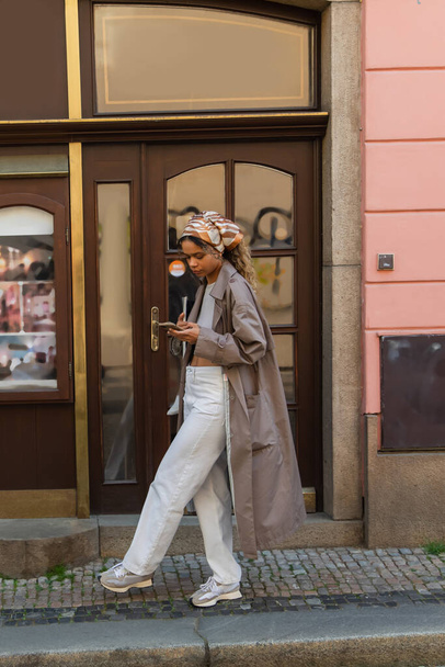 полная длина стильный африканский американец женщина в головном уборе и траншея пальто ходьба и использование смартфона в Праге  - Фото, изображение
