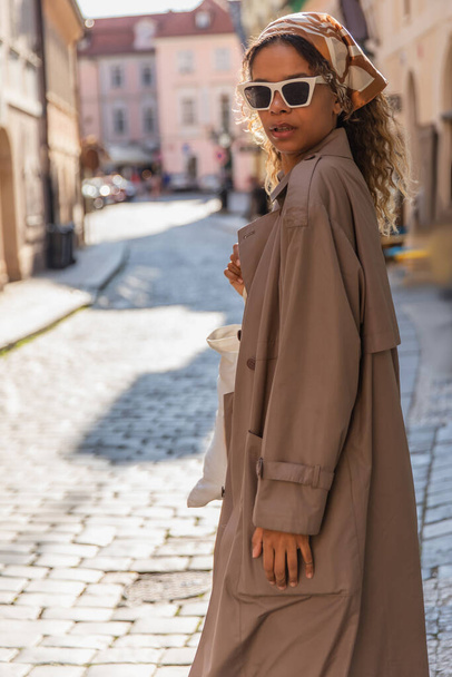 stylish african american woman in headscarf and sunglasses walking in trench coat on street in prague  - Foto, Imagen