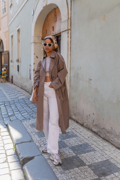 full length of stylish african american woman in headscarf and sunglasses posing in trench coat on street in prague  - Fotografie, Obrázek