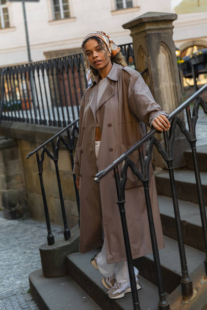 full length of trendy african american woman in trench coat standing on stairs  - 写真・画像