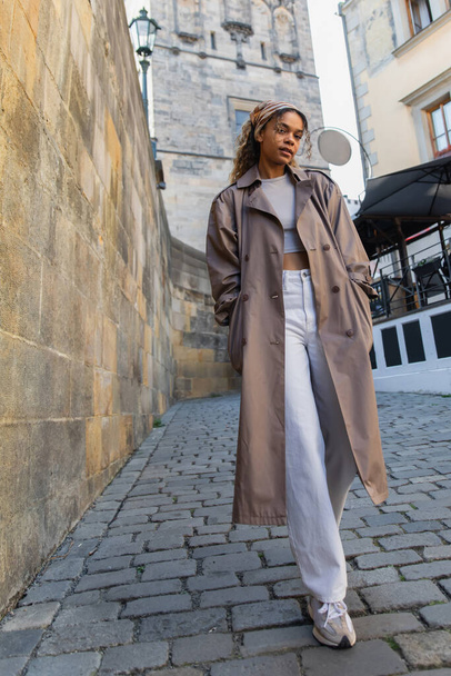 full length of stylish african american woman in trendy trench coat walking with hands in pockets near ancient wall in prague - Φωτογραφία, εικόνα