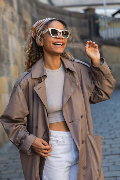 cheerful african american woman in trendy trench coat and sunglasses gesturing on street in prague - Fotó, kép
