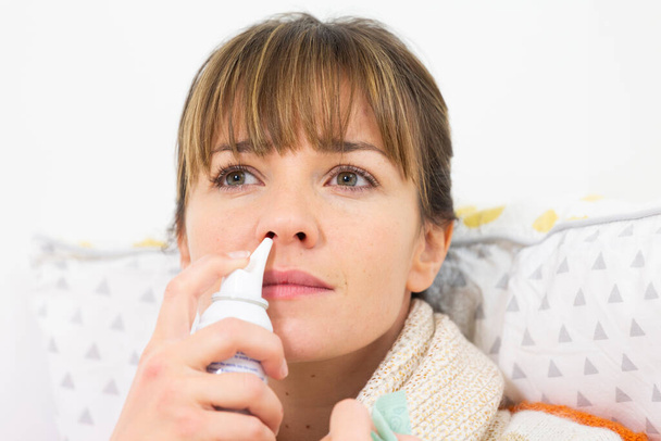 A young woman in bed suffering from a cold, with a nasal spray. - 写真・画像