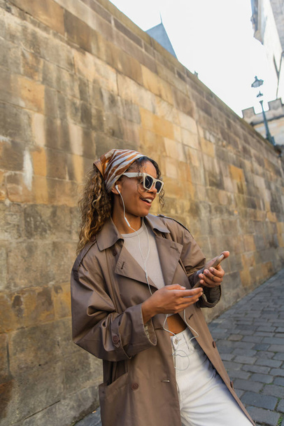 happy african american woman in trendy accessories listening music in wired earphones and using smartphone  - Fotó, kép