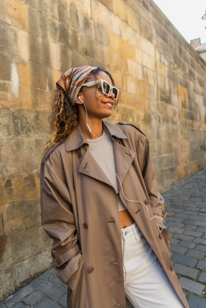 stylish african american woman in trendy accessories and trench coat listening music in wired earphones  - Fotó, kép