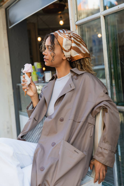 young african american woman in headscarf and stylish trench coat holding ice cream cone and sitting on chair in prague - Fotografie, Obrázek