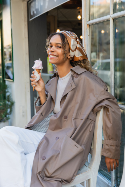 joyful african american woman in headscarf and stylish trench coat holding ice cream cone and sitting on chair in prague - Foto, Bild