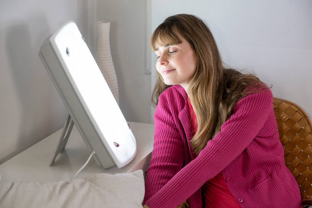 Woman sitting near a white light lamp for light therapy. - Photo, image