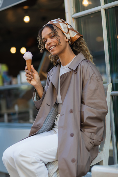 happy african american woman in headscarf holding ice cream cone and sitting on chair in prague - Foto, imagen