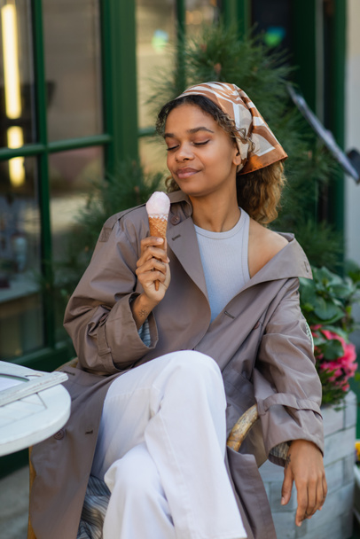 pleased african american woman in headscarf and stylish trench coat holding ice cream cone and sitting in cafe terrace  - Fotó, kép
