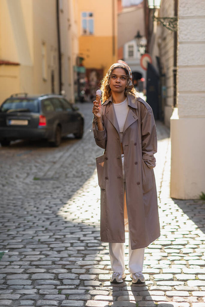 full length of dreamy african american woman in trench coat and headscarf holding ice cream cone on street in prague  - Foto, immagini