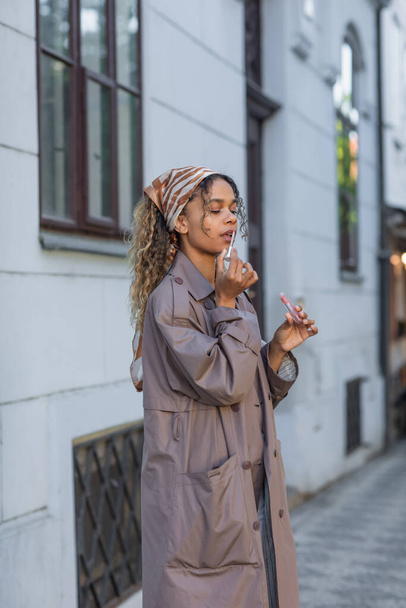 σγουρά αφροαμερικανή γυναίκα σε κομψό καμπαρντίνα και μαντίλα εφαρμογή lip gloss στο δρόμο στην Πράγα  - Φωτογραφία, εικόνα