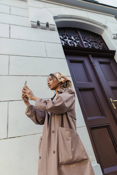 african american woman in trendy outfit and headscarf taking photo on smartphone near building in prague - Foto, Imagem