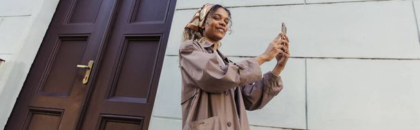 happy african american woman in trendy outfit and headscarf taking photo on smartphone near building in prague, banner - Valokuva, kuva