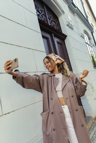 happy african american woman in trendy outfit taking selfie on smartphone near building in prague - Photo, image