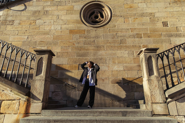 full length of stylish african american woman in oversize suit standing near ancient wall in prague - Foto, Imagem