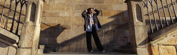 full length of stylish african american woman in oversize suit standing near ancient wall in prague, banner - Foto, Imagen
