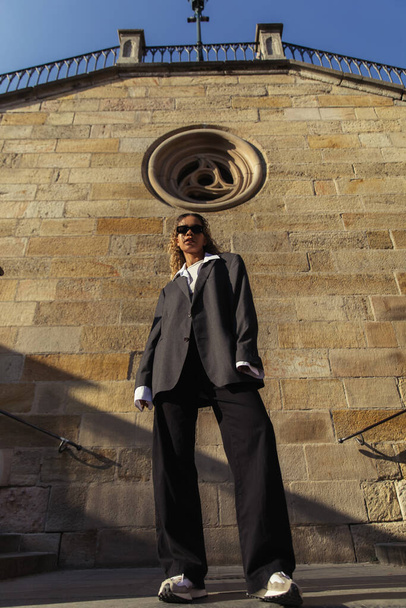 low angle view of trendy african american woman in sunglasses and oversize suit standing near ancient wall on street in prague - Fotoğraf, Görsel