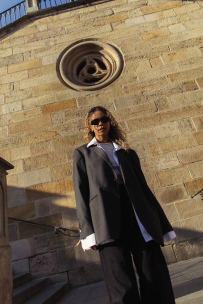 low angle view of stylish african american woman in sunglasses and oversize suit standing near ancient wall on street in prague - Foto, imagen