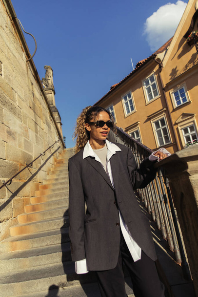 stylish african american woman in sunglasses and oversize suit on stairs of ancient street in prague  - Fotó, kép