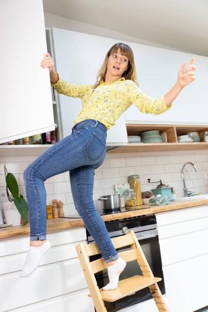 Young woman in her kitchen falling from a chair. - Фото, изображение