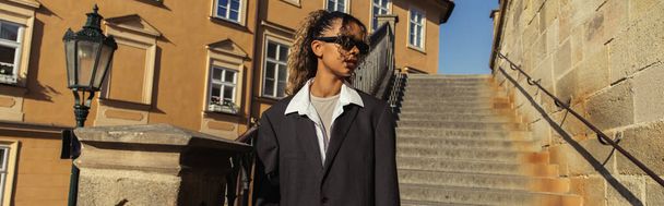 stylish african american woman in sunglasses and oversize suit standing near stairs on street in prague, banner - Foto, Bild