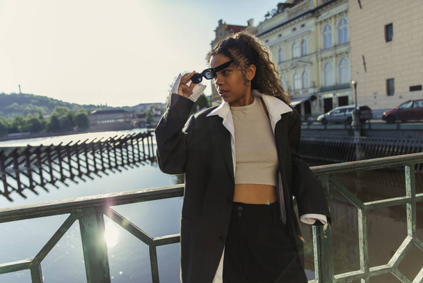 stylish african american woman in oversize suit wearing sunglasses and standing near river in prague  - Foto, Imagen