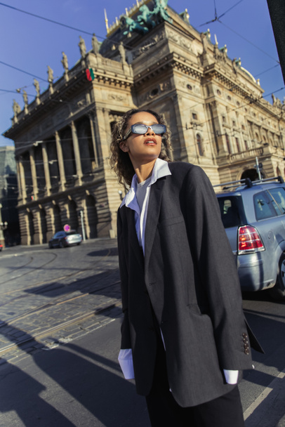 κομψή Αφροαμερικανή γυναίκα με μεγάλο σακάκι και γυαλιά ηλίου που στέκεται κοντά στο εθνικό θέατρο στην Πράγα - Φωτογραφία, εικόνα