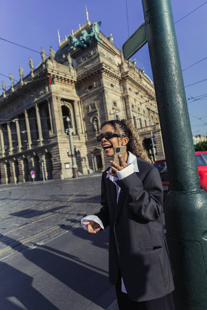 happy african american woman in oversized blazer and sunglasses holding smartphone near national theatre in prague - Photo, Image